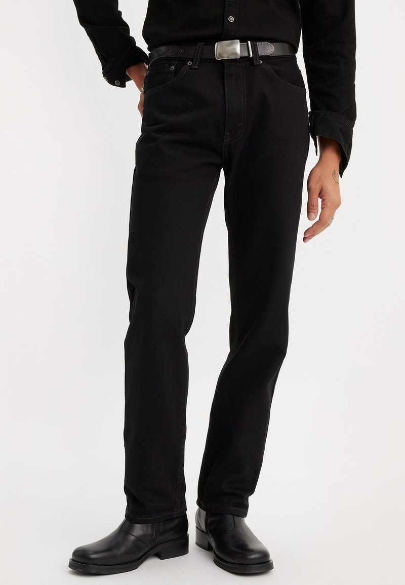 Moški nosi črne ravne hlače, črn pas s srebrno zaponko, črno dolgotrajno majico in črne usnjene gležnjarje.