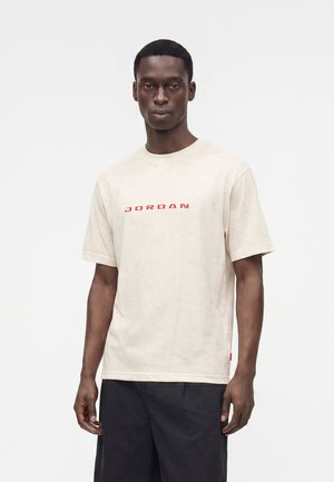 Mężczyzna ubrany w beżowy T-shirt z czerwonym tekstem "JORDAN" i czarne spodnie, stojący na jasnym, jednolitym tle.