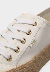 Baskets en toile blanche avec des lacets blancs passant par des œillets dorés et une semelle en jute tissée, avec une petite étiquette en tissu "ONLY".