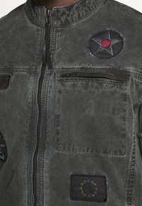 Harmaa, teksturoitu takki, jossa on korkea kaulus ja etuvetoketju. Siinä on kolme merkkiä: punainen tähti ja kaksi lippua. Useita taskuja näkyvissä.