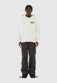 Weißer Hoodie mit einer Vorderentasche und schwarzem Grafikdesign, kombiniert mit locker sitzenden schwarzen Cargo-Hosen, die mehrere Taschen und einen Kordelzug haben.