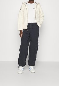 Persona che indossa una giacca di pile bianca con zip, una maglietta bianca, pantaloni cargo neri larghi e sneakers bianche con dettagli blu, in piedi su uno sfondo neutro.
