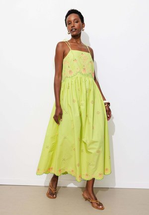 Femme portant une robe sans manches vert citron avec broderies florales et ourlet festonné, debout contre un mur blanc, des sandales marron et des boucles d'oreilles créoles.