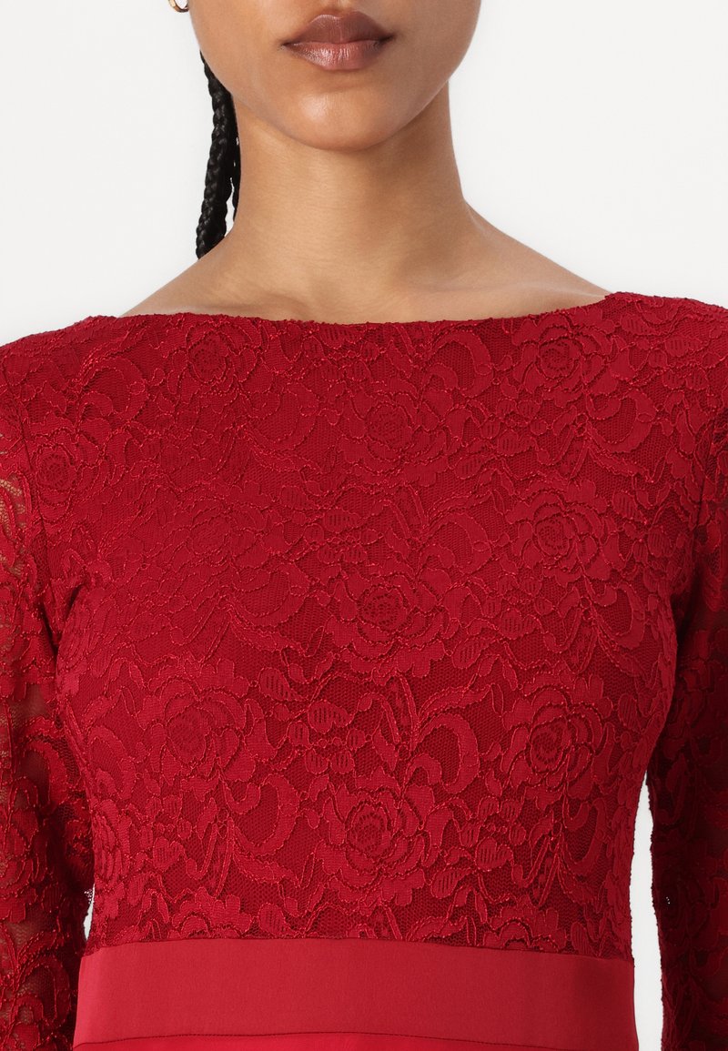 Rotes Spitzenoberteil mit floralem Muster, Boot-Ausschnitt und langen Spitzenärmeln. Glattes rotes Stoffdetail am Taillenrock.