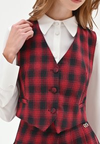 Gilet a quadri rosso e nero dal design aderente, con camicia bianca a collo sotto, tre bottoni e un dettaglio con logo.