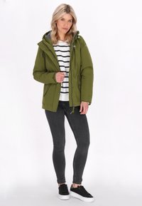 Olivgrön huva-jacka med dragkedja och tryckknappar, grått foder, lapplaggslommer, bärs över en vit och svart randig topp med svarta skinny jeans.