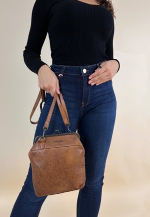 Femme portant un haut noir à manches longues et un jean bleu foncé, tenant un sac à main en cuir marron par ses anses à son côté.