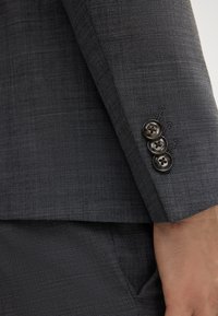 Grauer Stoffblazer mit dezentem Muster und tailliertem Schnitt. Drei dunkle Knöpfe an den Ärmeln unterstreichen das Design.