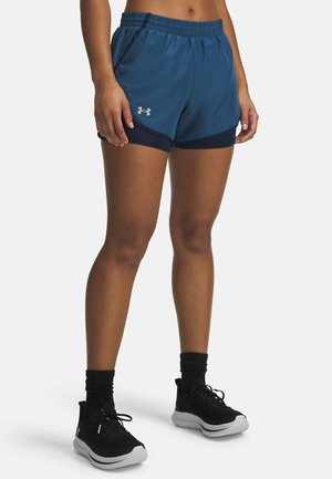 Blue athletic shorts with an elastic waistband, feature a contrasting dark blue trim, and side pockets. Worn with black sneakers and socks.