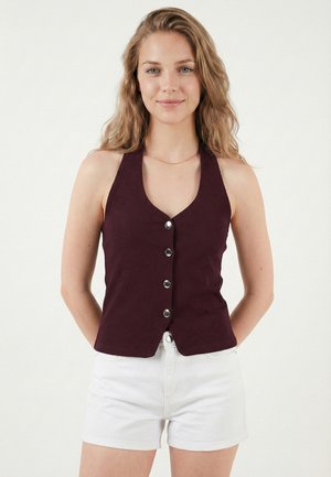 Młoda kobieta z długimi falującymi włosami, ubrana w bezrękawnik w ciemnym fioletowym kolorze z guzikami oraz białe szorty, stoi na jednolitym tle.