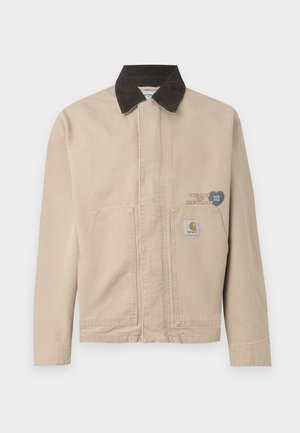 Beige canvas jacket with a black corduroy collar, featuring a patch on the left chest and two front pockets. Simple, structured design.
