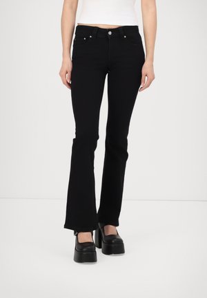 Femme portant un jean évasé noir, un haut court blanc et des chaussures compensées noires, debout devant un fond blanc uni.
