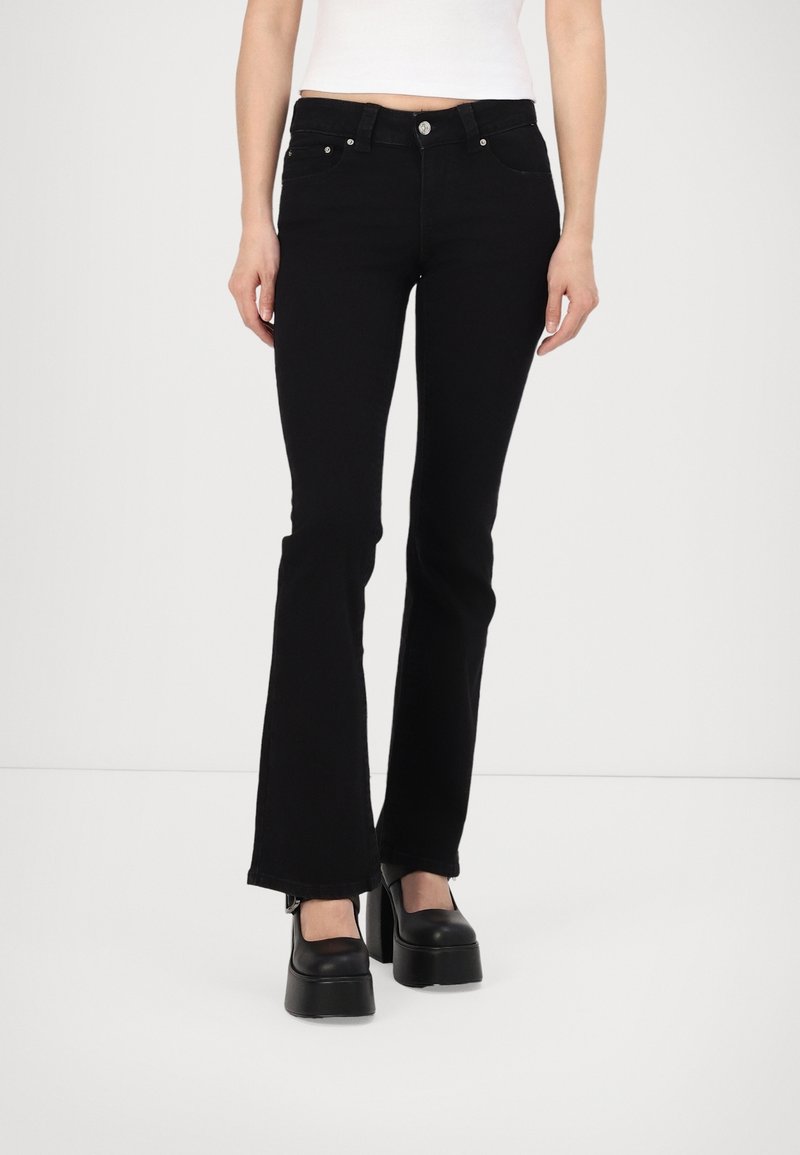 Femme portant un jean évasé noir, un haut court blanc et des chaussures compensées noires, debout devant un fond blanc uni.