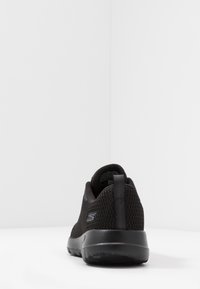 Sort atletisk sko med et mesh-overdel, rund hæl og slip-on design. Har en tekstureret overflade og en logodetalje på siden.