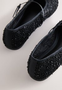 Paire de ballerines noires à bout rond, recouvertes de petites perles et sequins noirs, avec fines brides à la cheville.