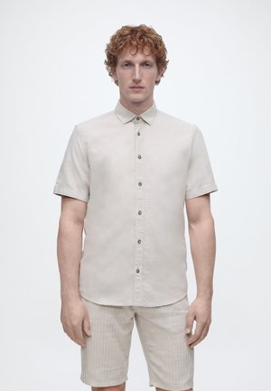 Homme aux cheveux roux bouclés portant une chemise à manches courtes beige clair et un short beige rayé sur un fond blanc uni.