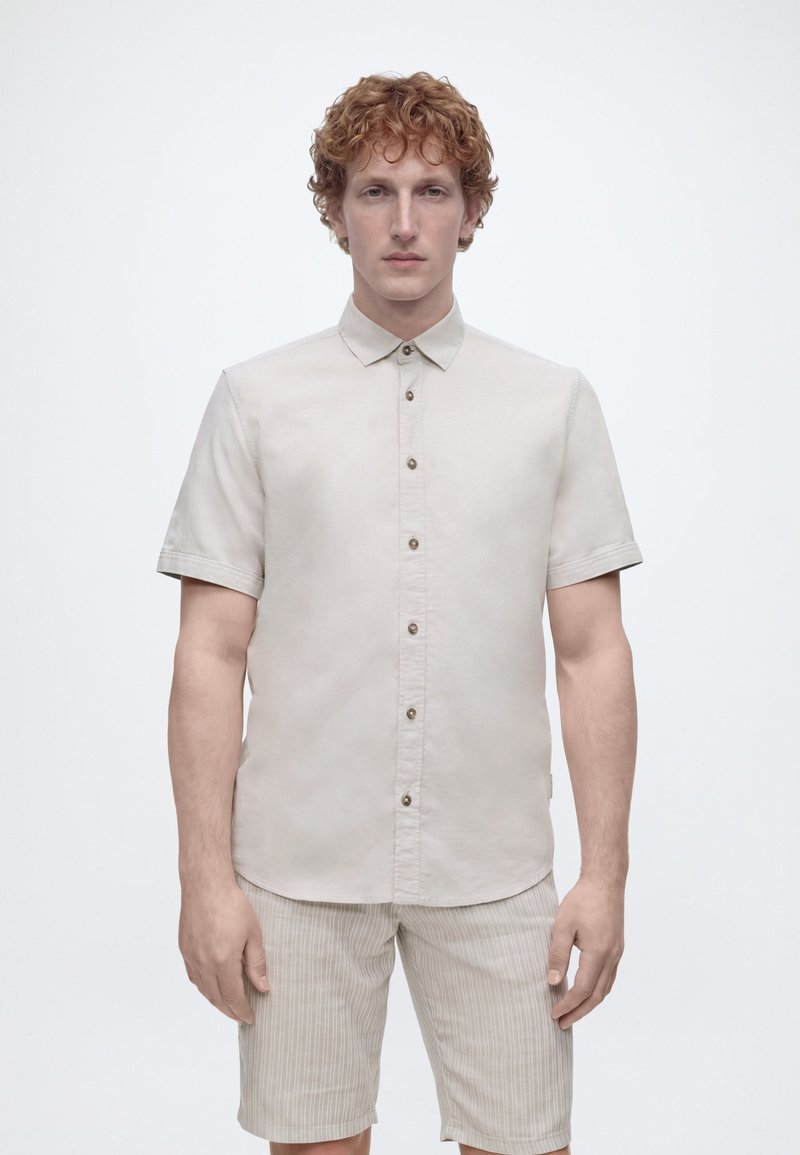 Άνδρας με μπούκλια κόκκινα μαλλιά φοράει ανοιχτό μπεζ πουκάμισο με κοντά μανίκια και μπεζ ριγέ σορτς, σε απλό λευκό φόντο.