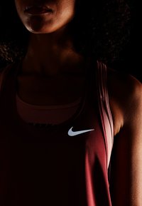 Bordowy sportowy top na ramiączkach wykonany z gładkiej tkaniny, z białym logo Nike na piersi i warstwowym krojem z wycięciem typu racerback.