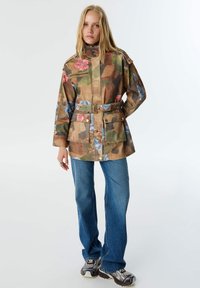 Bunda s camo vzorem a květinovým potiskem, vysokým límcem, páskem v pase a dvěma předními kapsami. Spárována s modrými džíny a vzorovanými teniskami.