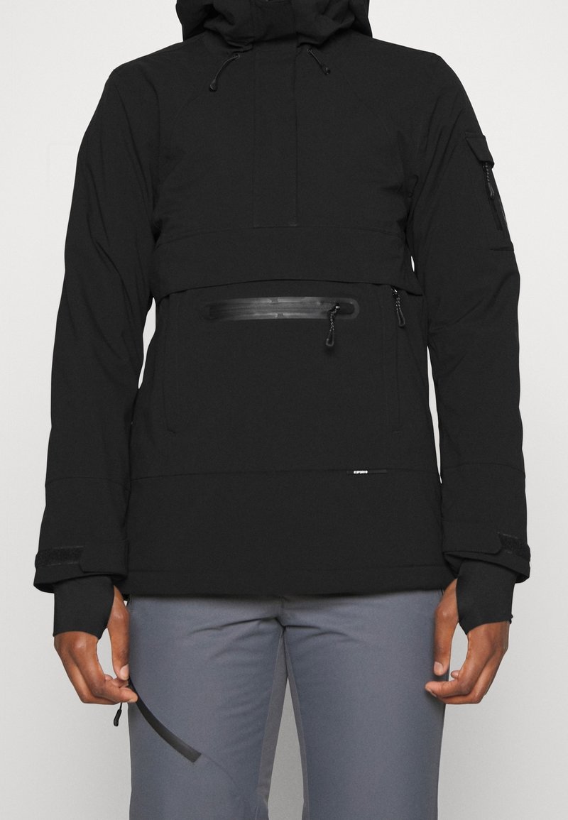 Veste noire à capuche avec poche zippée à l'avant, manches longues avec trous pour les pouces, et pantalon gris sur une personne debout.