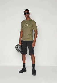 Camisa de manga curta verde oliva com logotipo, calções pretos, sapatos pretos e óculos de sol. Segurando um capacete cinza, em pé contra um fundo neutro.
