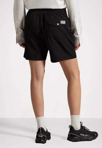 Schwarze Shorts mit einem elastischen Bund, einer Gesäßtasche mit einem Etikett und einer glatten Stofftextur. Getragen mit schwarzen Sneakers und weißen Socken.