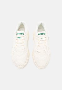 Baskets Lacoste en tissu blanc cassé avec des motifs texturés, arborant un logo vert sur la languette et un design de semelle épaisse.