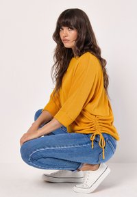 Gul rippad tröja med halv ärm, med samlade detaljer på sidorna. Matchad med blå denimjeans och vita sneakers.