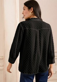 Blusa nera con motivo a pois testurizzati, maniche lunghe e scollo arricciato. Presenta spalle arricciate e una vestibilità rilassata. Indossata con jeans.