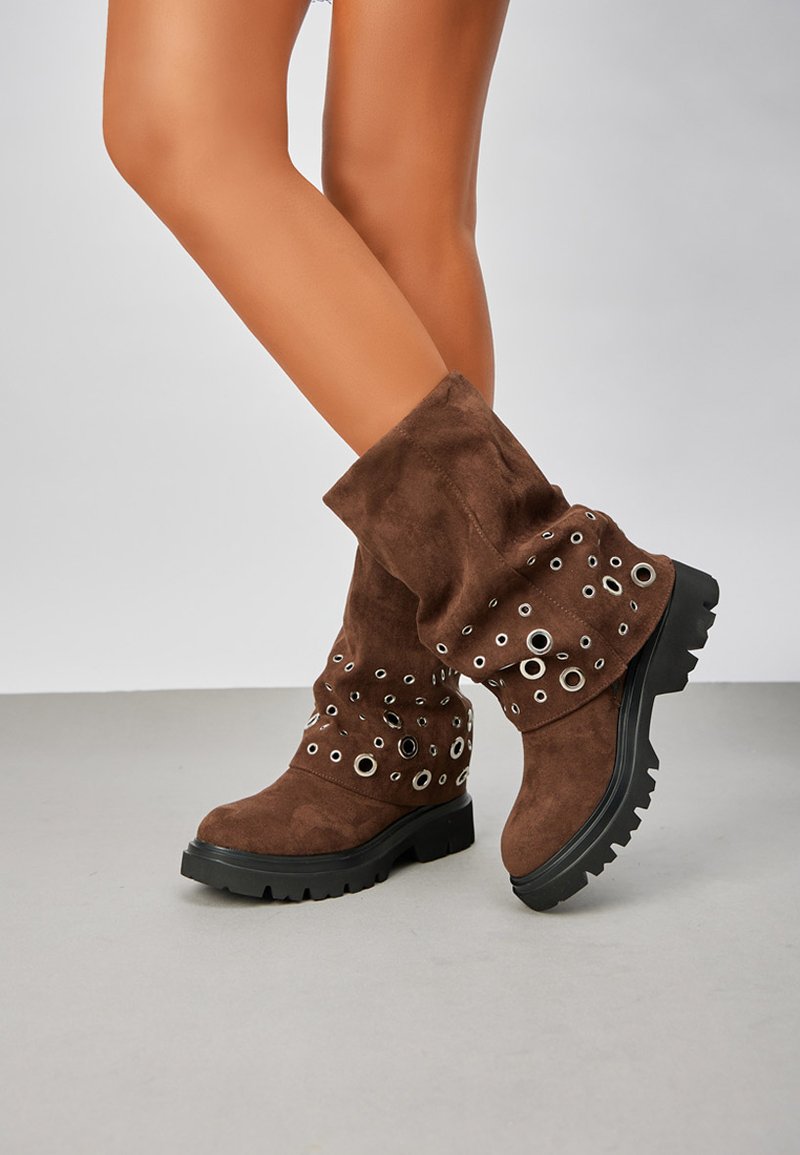 Bottes en daim marron mi-mollet avec semelles épaisses noires et détails en œillets argentés, portées sur des jambes croisées devant un fond uni.