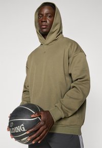 Jeune homme portant un sweat à capuche olive tenant un ballon de basket Spalding noir devant un fond clair uni.