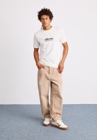 Fehér pamut póló „ellesse since 1959” nyomtatott logóval, bézs, bő szabású nadrággal és fehér sportcipővel, barna talppal és sötétkék részletekkel.