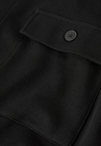 Tissu noir avec un tissage texturé, comprenant un détail de poche boutonnée. La poche possède un rabat et un bouton rond à quatre trous.