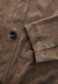 Gros plan sur une veste en daim marron montrant une manche retroussée, un bouton noir et un détail de poche cousu.