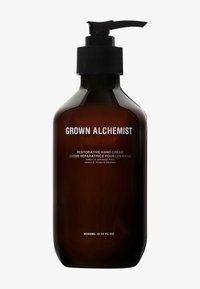 Bouteille en verre ambre foncé avec un bouchon-pompe noir. L'étiquette présente un texte blanc indiquant "Crème pour les mains réparatrice Grown Alchemist." 300ml.
