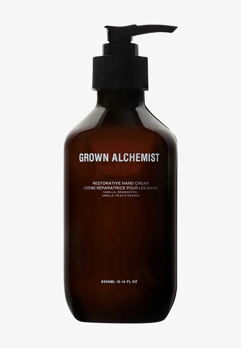 Bouteille en verre ambre foncé avec un bouchon-pompe noir. L'étiquette présente un texte blanc indiquant "Crème pour les mains réparatrice Grown Alchemist." 300ml.
