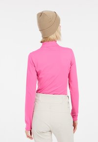 Maglietta aderente a maniche lunghe di un vivace colore rosa, realizzata in tessuto elastico, con colletto alto e cuciture piatte. Indossata con pantaloni chiari e un berretto beige.