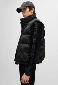 Black puffer vest featuring a high collar, quilted design, and a logo patch. Accented with black velvet sleeves and patterned side details.
