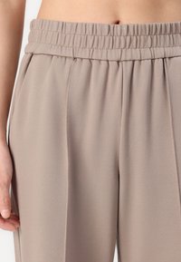 Světlé béžové kalhoty s elastickým pasem, rovnými nohavicemi a jemnou, hladkou tkaninou; obsahují detail boční kapsy.