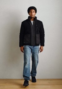 Giacca in pile nera con dettagli grigi, abbinata a jeans in denim azzurro chiaro e sneakers nere. Modello in piedi su un pavimento di legno.