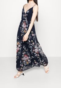 Kvinna som går i en marinblå blommig maxiklänning med spaghetti-remmar och vita klacksandaler med remmar på en enkel vit bakgrund.