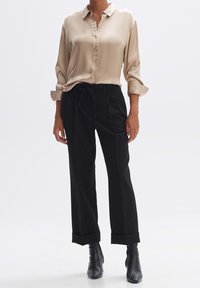 Chemise beige en satin à boutons avec manches longues, associée à un pantalon noir ample avec taille à cordon et revers roulés. Bottines noires.
