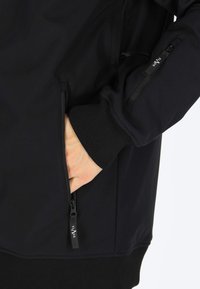 Czarna kurtka softshell z zamykanym na zamek bocznym kieszonką na lewym rękawie i ręką w kieszeni bocznej. Posiada ściągacze przy mankietach oraz gładką teksturę.