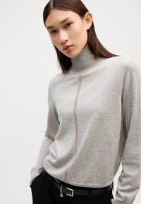 Gai pelēks turtlenecks džemperis mīkstā audumā ar šuvju detaļu pa centru, saskaņots ar melnām biksēm un jostu ar zelta akcentiem.