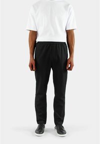 Svart elastisk bukse med smal passform og jevn tekstur, sammen med en enkel hvit t-skjorte med korte ermer og svarte joggesko.