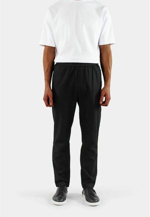 Schwarze elastische Hose mit schmaler Passform und glatter Textur, kombiniert mit einem schlichten weißen T-Shirt mit kurzen Ärmeln und schwarzen Sneakern.