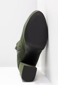 Botín de ante verde con tacón cuadrado, detalle de cremallera lateral y suela negra estriada. Textura suave con un acabado mate.