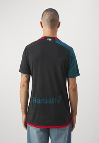 Camiseta deportiva negra con ribete rojo y acentos en teal, que presenta un gran logotipo azul de "Herbalife" en la parte posterior. La tela parece suave y ligera.