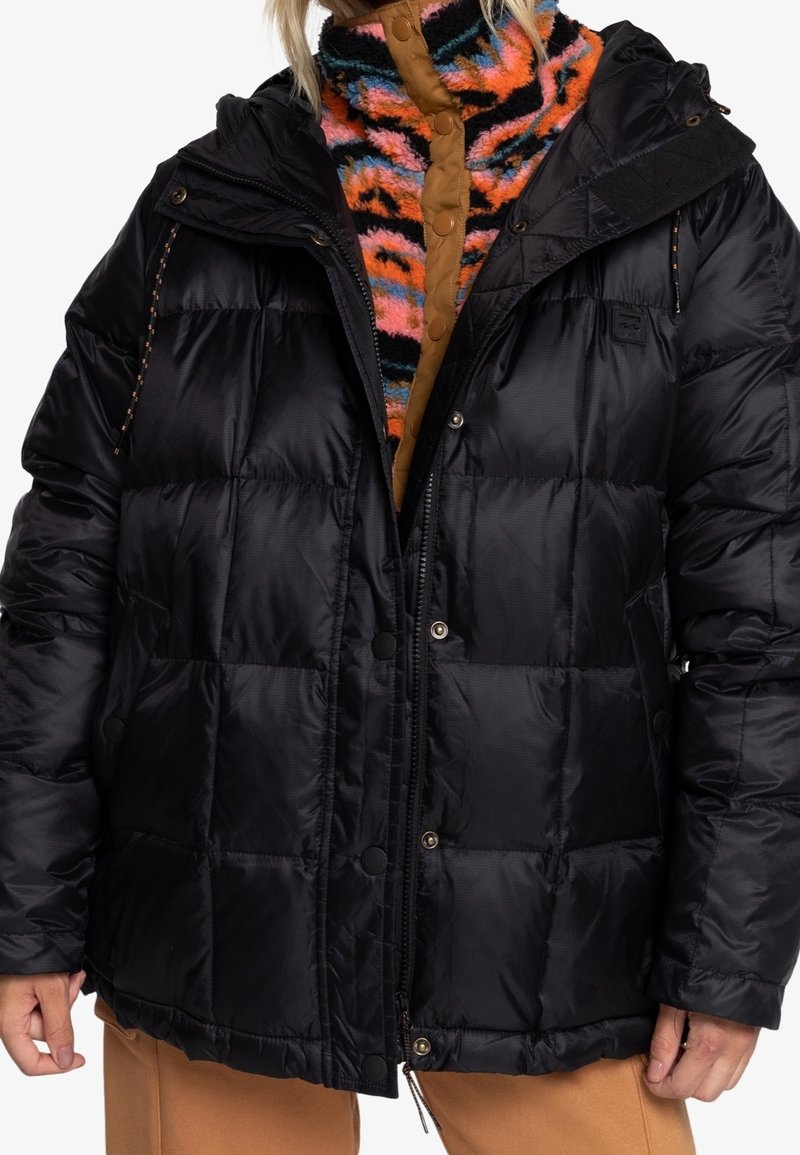 Veste matelassée noire à manches longues portée sur une veste colorée à rayures tigre en tissu duveteux avec boutons, et pantalon marron, portée par une personne aux cheveux blonds.