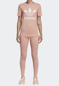Person som bär en matchande outfit i blek rosa från Adidas med en t-shirt med logo, leggings med vita ränder och rosa sneakers, stående rakt mot en vit bakgrund.